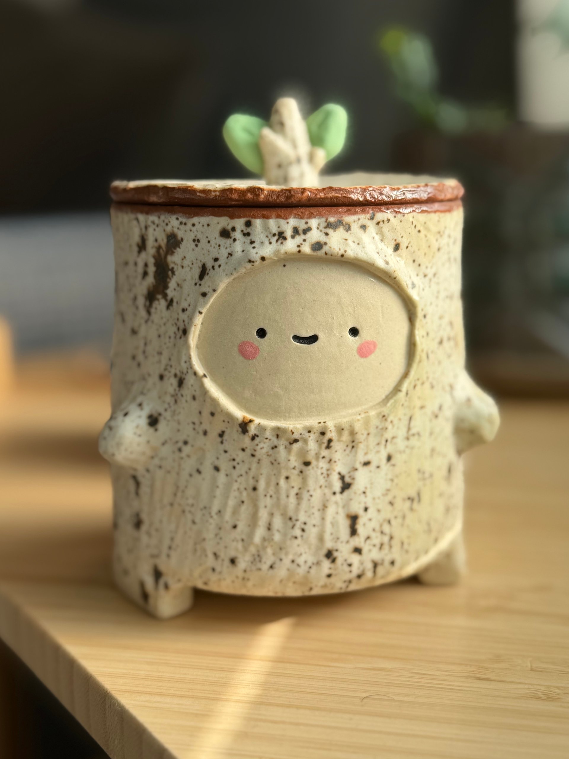 A lidded jar shaped like a birch stump, speckled cream and dark, with a face and green sprouts on the lid