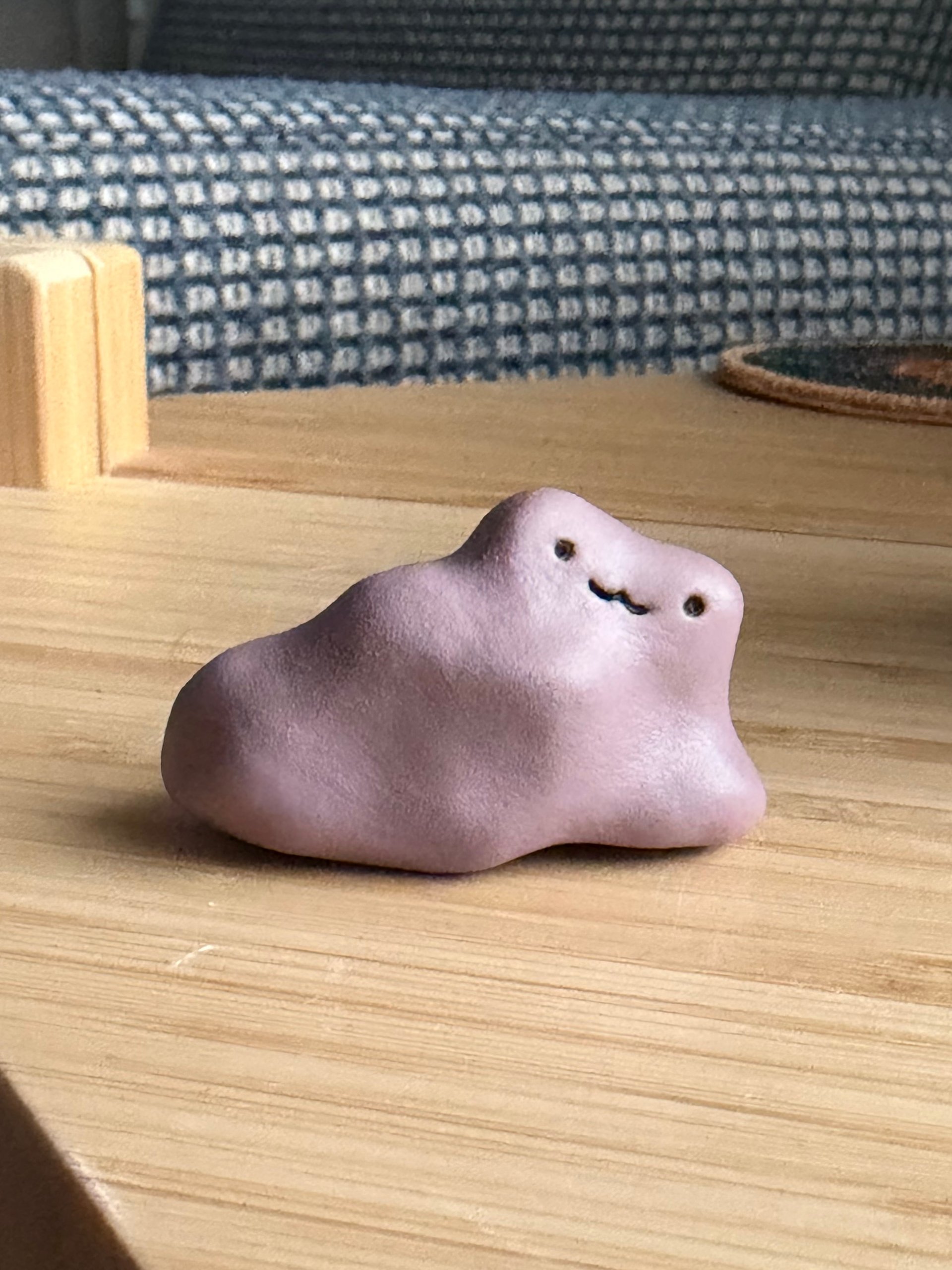 A small pale lilac clay frog with a simple smiling face, sitting on a wooden table