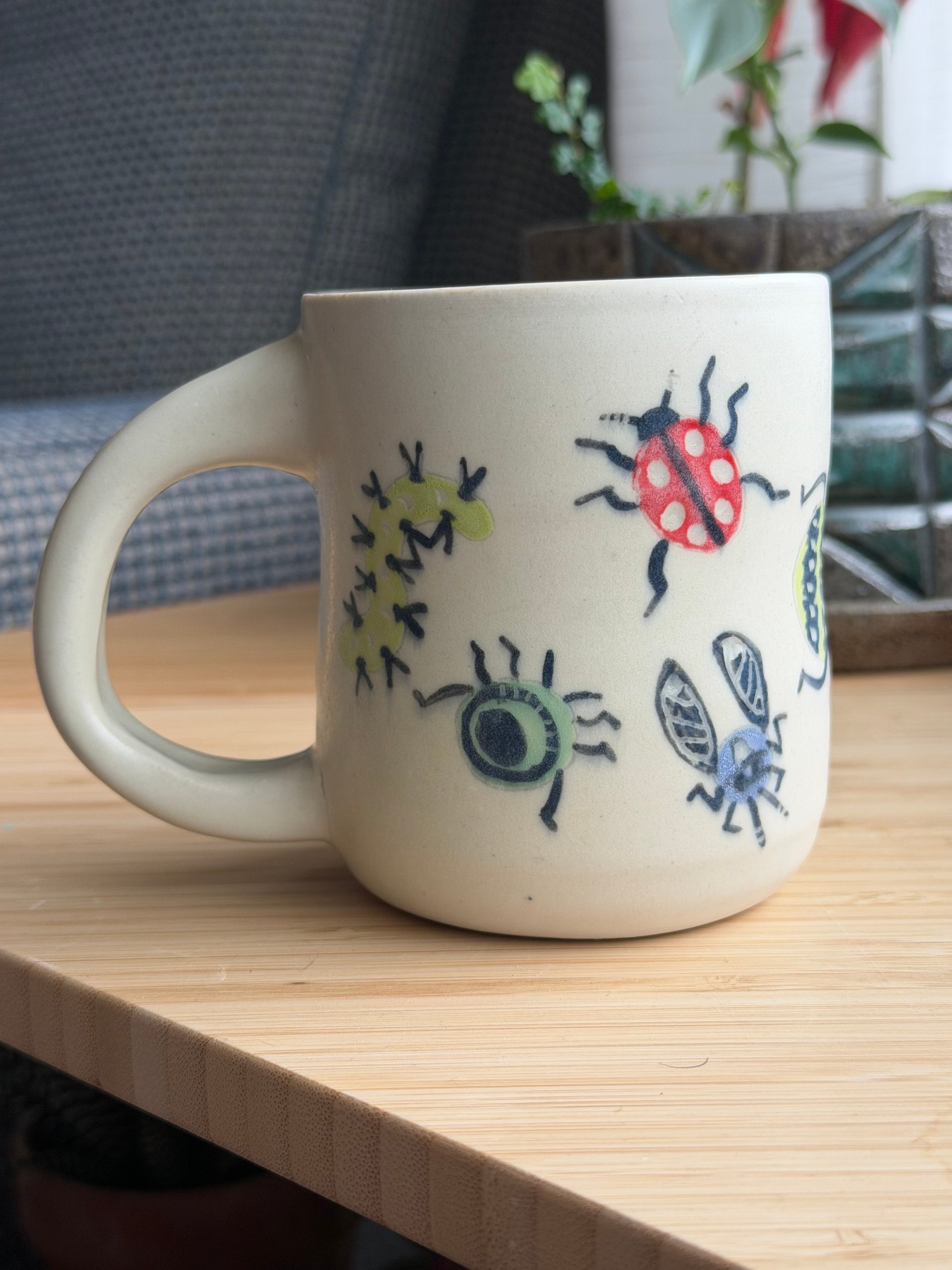 A cream mug painted with small bugs, its handle to the left and a red ladybug near the rim