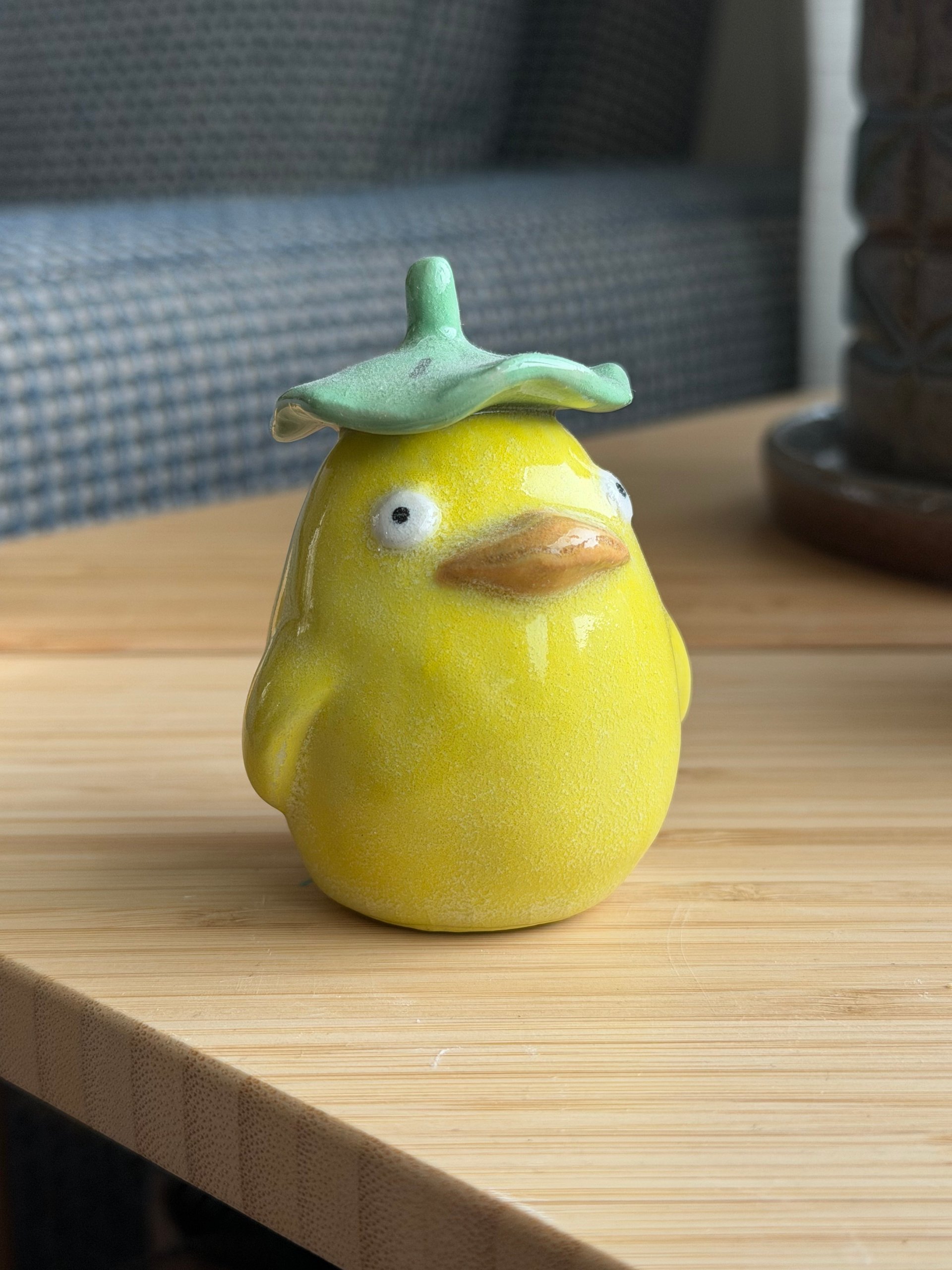A round yellow ceramic duck wearing a green lilypad as a hat, sitting on a wooden table