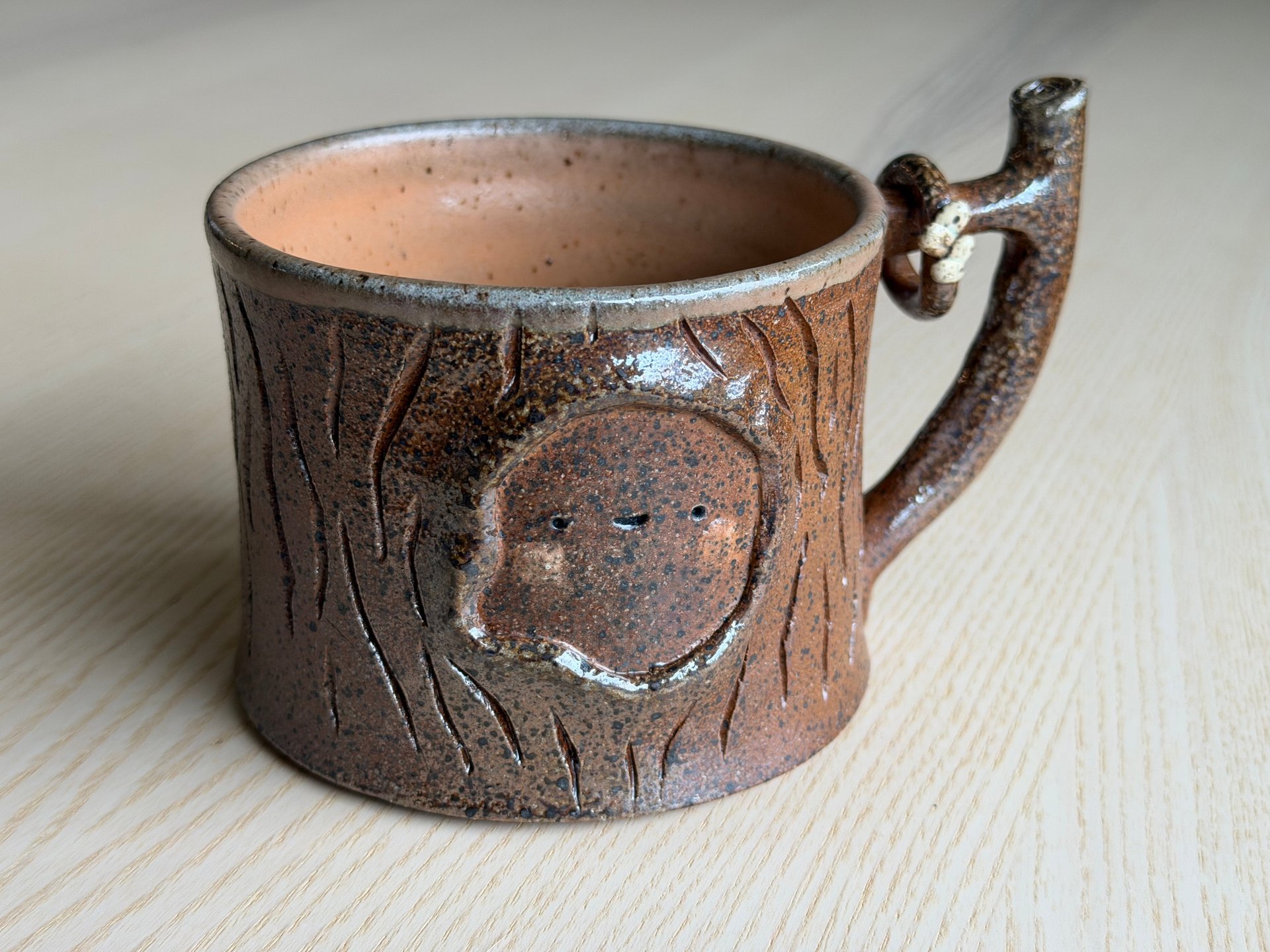 A dark brown speckled mug with carved bark and a branch-shaped handle