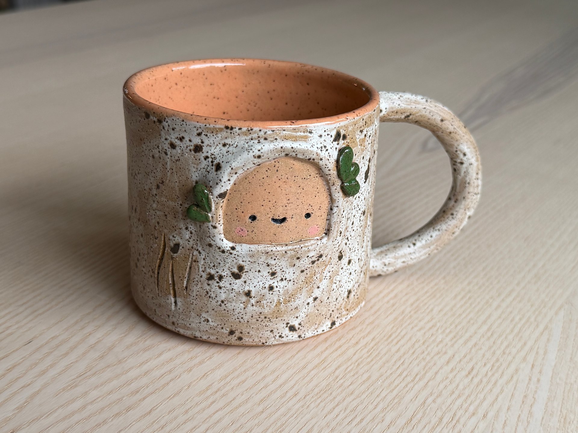 A pale speckled mug with green ivy leaves and a rosy-cheeked face